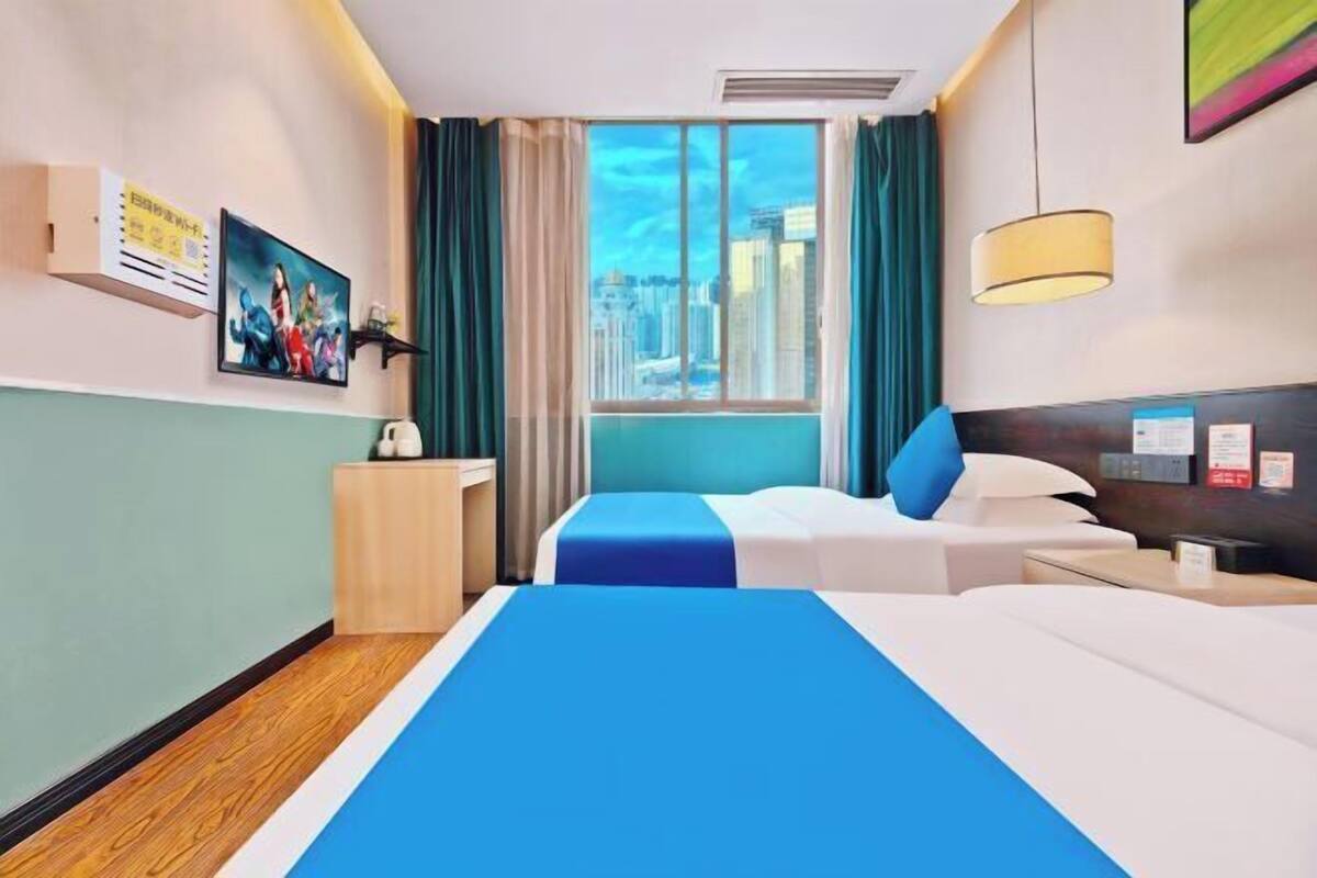 Ein Hotelzimmer mit einem großen Bett, einem Einzelbett, einem Schreibtisch, einem Sessel, einem Fenster mit Vorhängen und einem Bild an der Wand.