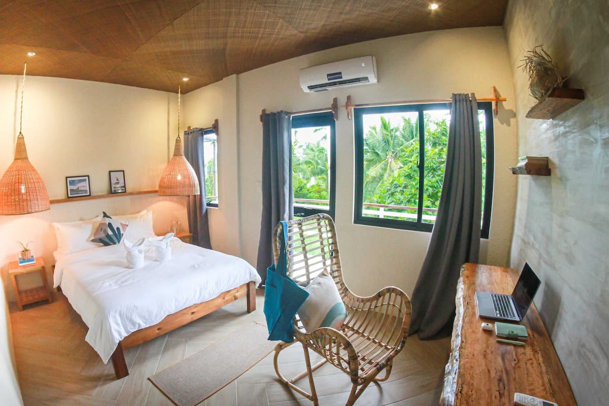 A bedroom with a bed, bedside table, lamp, chair, laptop, and an air conditioning unit.