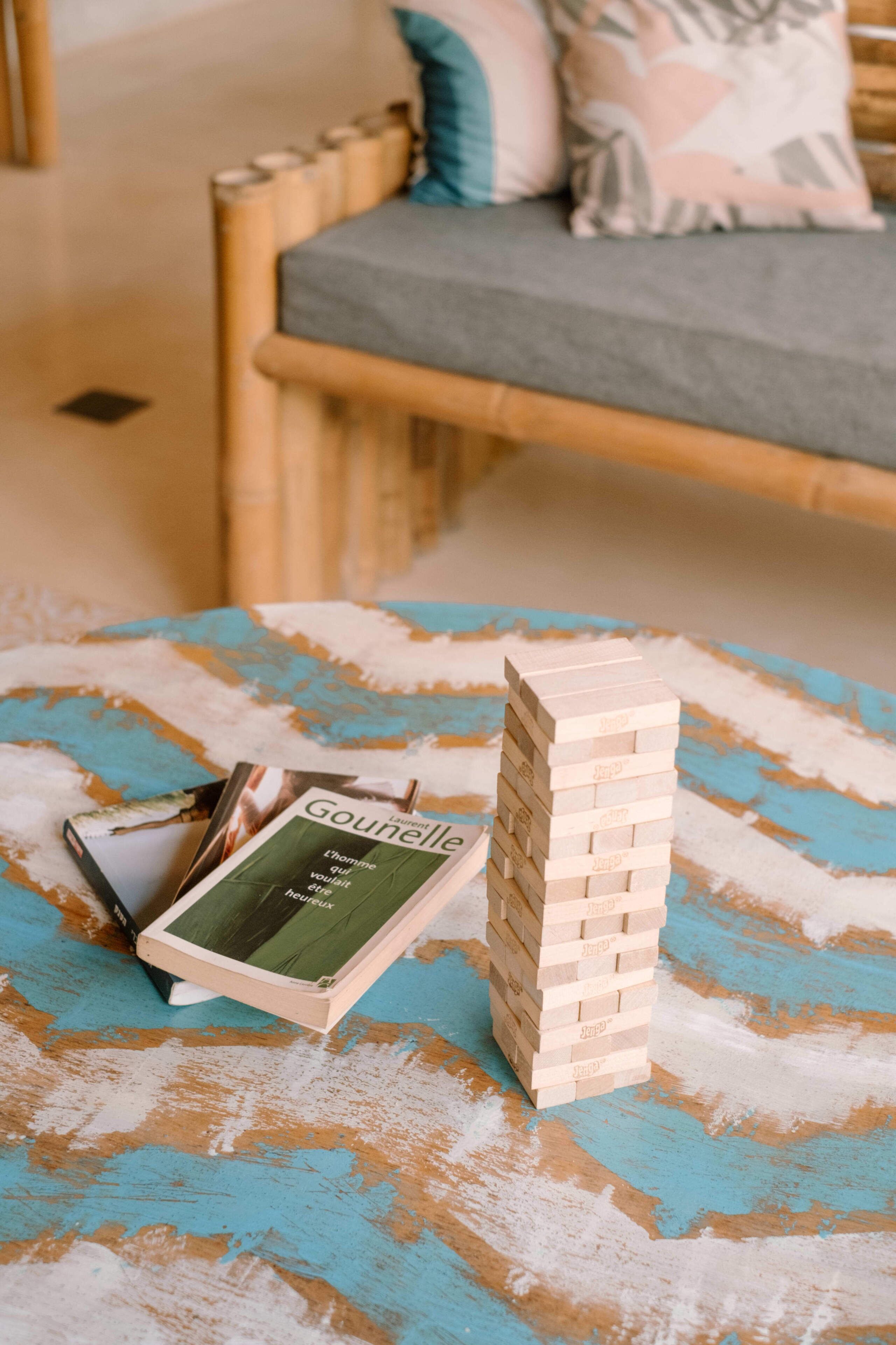 A wooden bed frame with a grey mattress and patterned pillows, a blue and orange textured table with a stack of wooden blocks and a book titled 'Gounelle.'