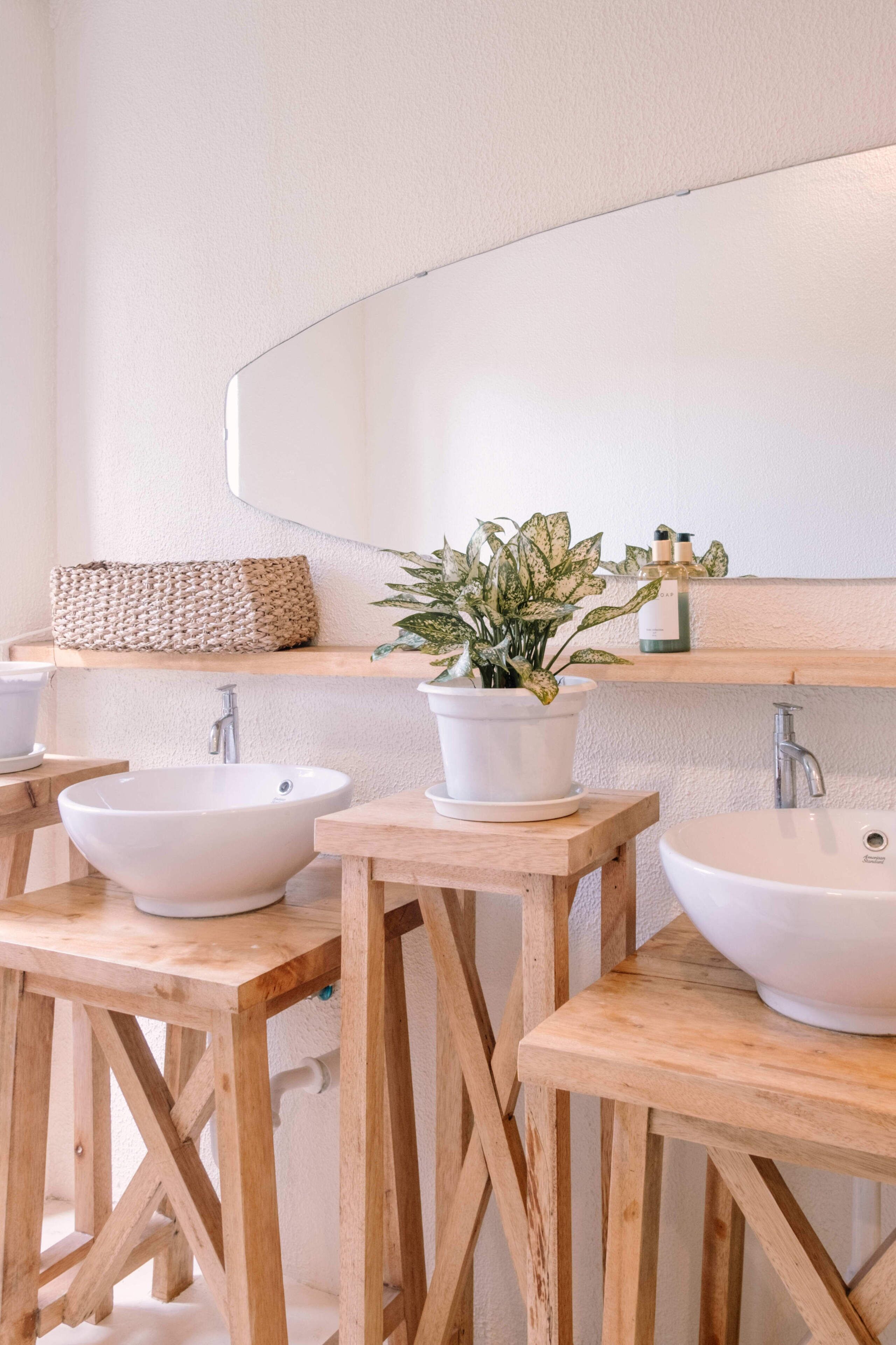 Une salle de bain avec deux lavabos blancs, des tabourets en bois, un miroir, un panier en osier et une plante en pot.