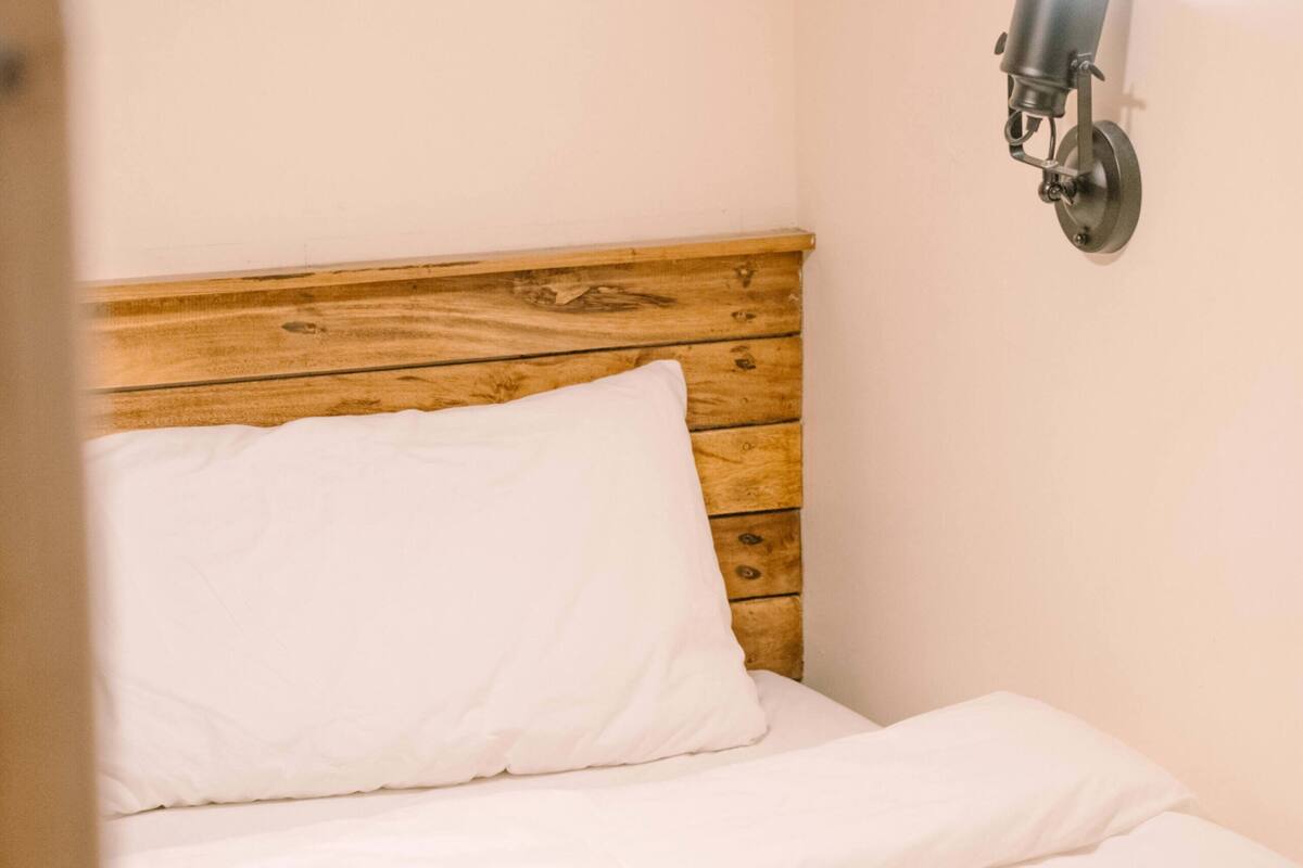A neatly made bed with white linens and a rolled towel, a wooden headboard, and a wall-mounted light fixture.