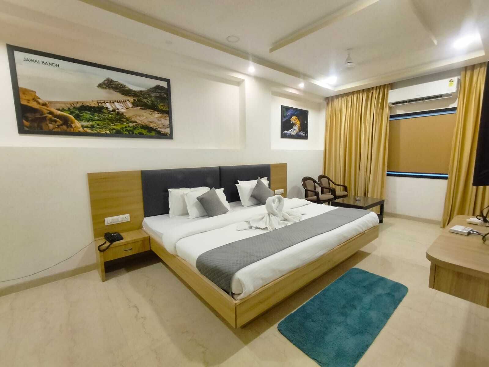 A hotel room with a large bed, a desk, a chair, a nightstand, a wall-mounted air conditioner, and two framed pictures on the wall.