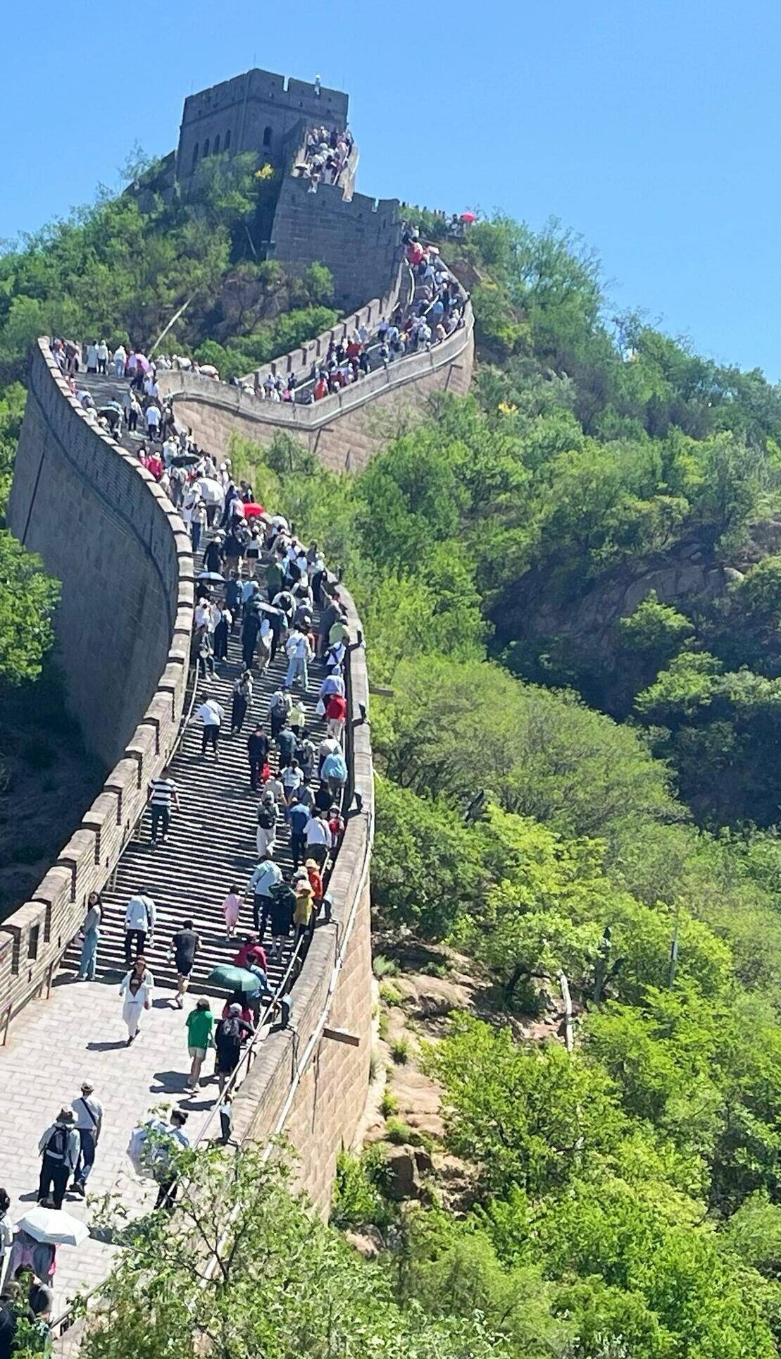 A crowded walkway on a steep, curved stone wall with a watchtower at the top.
