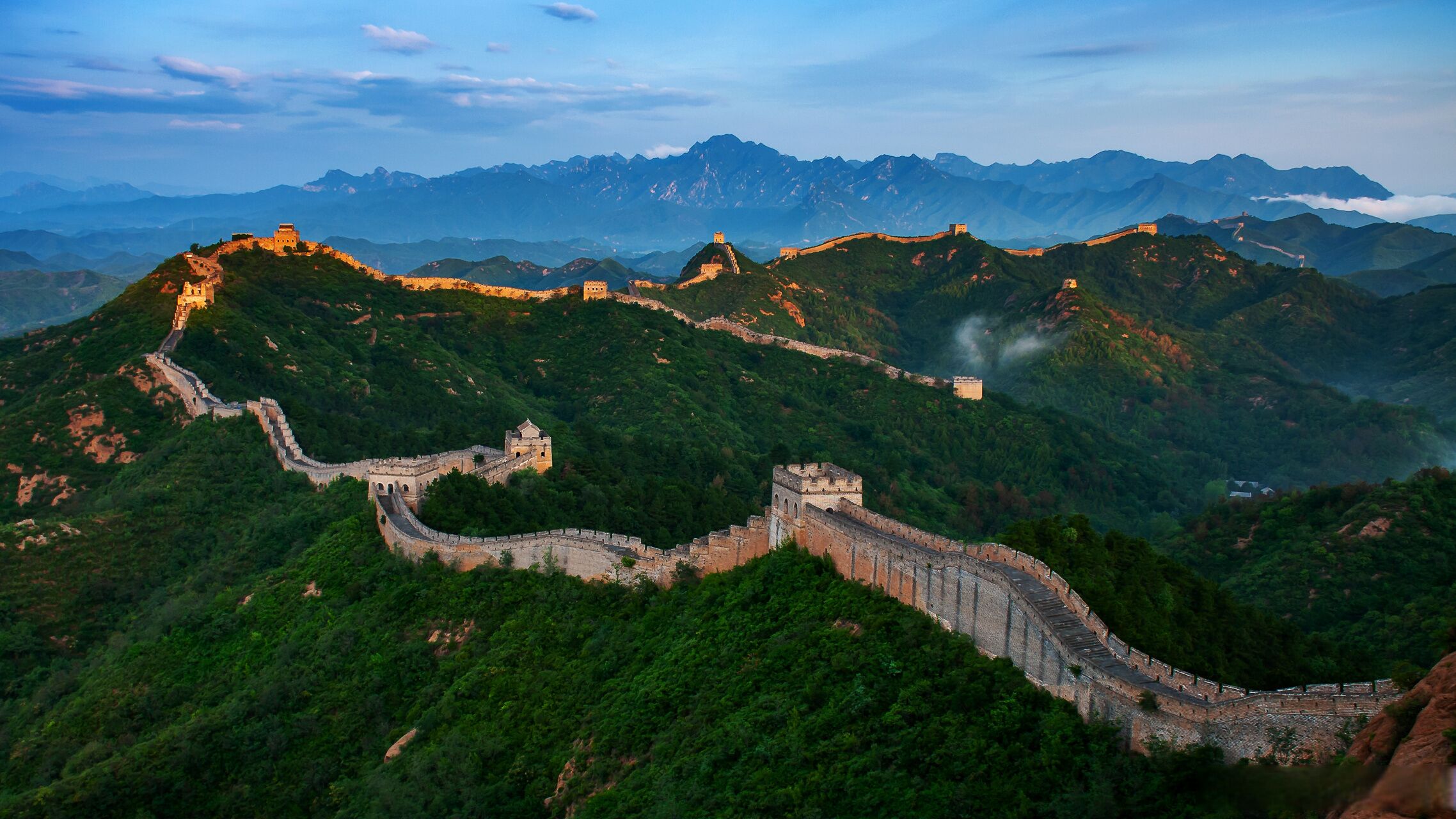 De Chinese Muur kronkelend door glooiende groene heuvels, met een bergachtig landschap op de achtergrond.