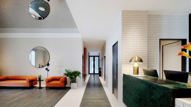 Un hall d’hôtel moderne avec un comptoir d’accueil, un long couloir et un espace salon agrémenté de canapés orange.