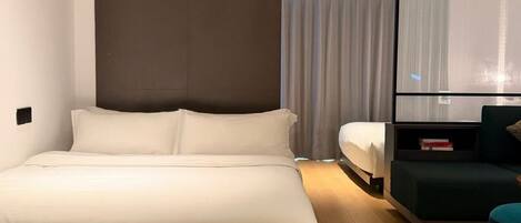 A modern hotel room with a large bed, a seating area, and a large window with curtains.