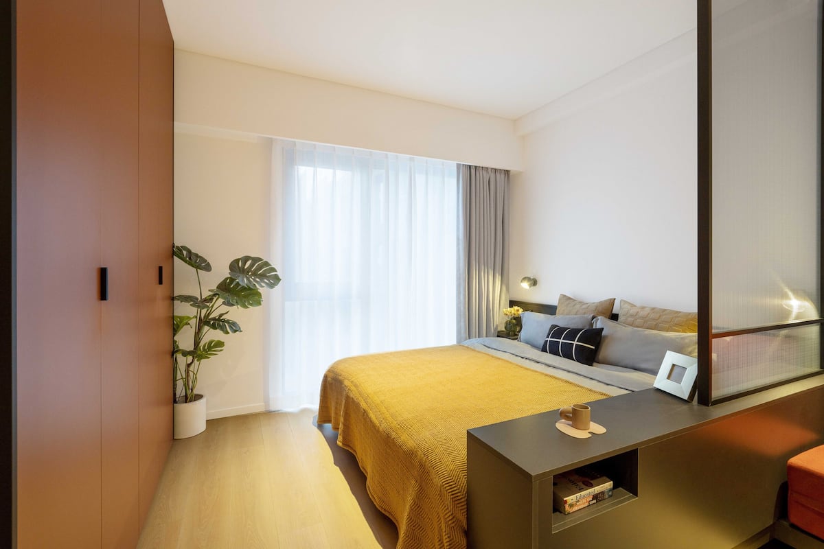 A modern hotel room with a large bed, a bedside table, a potted plant, and a large window with curtains.