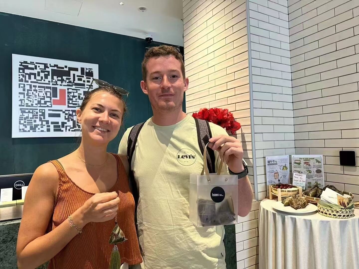 A man and a woman standing in a room with a white brick wall, a table with food items, and a QR code on the wall.