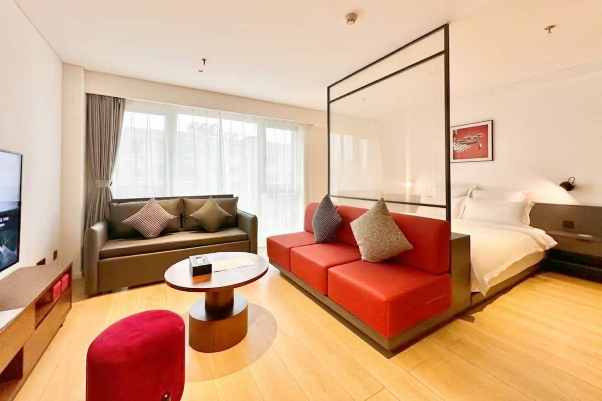 A modern hotel room with a sofa, a red couch, a bed, a TV, and a large window with curtains.