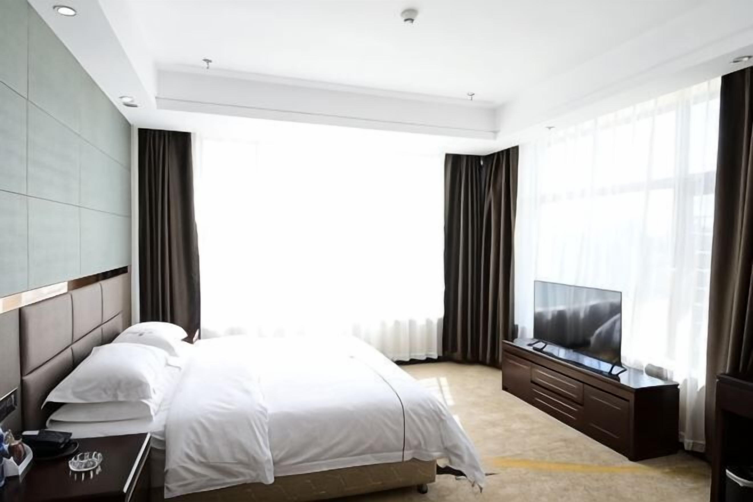 A hotel room with a large bed, a TV, a seating area with a sofa, and a large window with curtains.