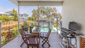 Un balcón con mobiliario de madera, una parrilla y vistas a una piscina y casas vecinas.