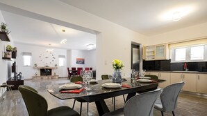 A modern dining area with a long table set for six, surrounded by chairs. A fireplace is visible in the background, and the room has a high ceiling with recessed lighting. - Villa panagiotis (Κίσσαμος)