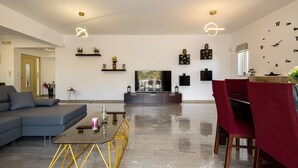 A modern living room with a grey sectional sofa, a glass coffee table, a TV on a dark stand, and a dining area with red upholstered chairs. - Villa panagiotis (Κίσσαμος)