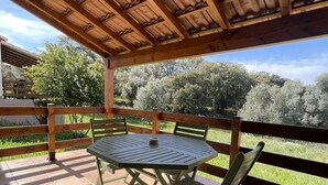 Une terrasse couverte avec une table ronde et des chaises, des rampes en bois et une vue sur les arbres et les collines.