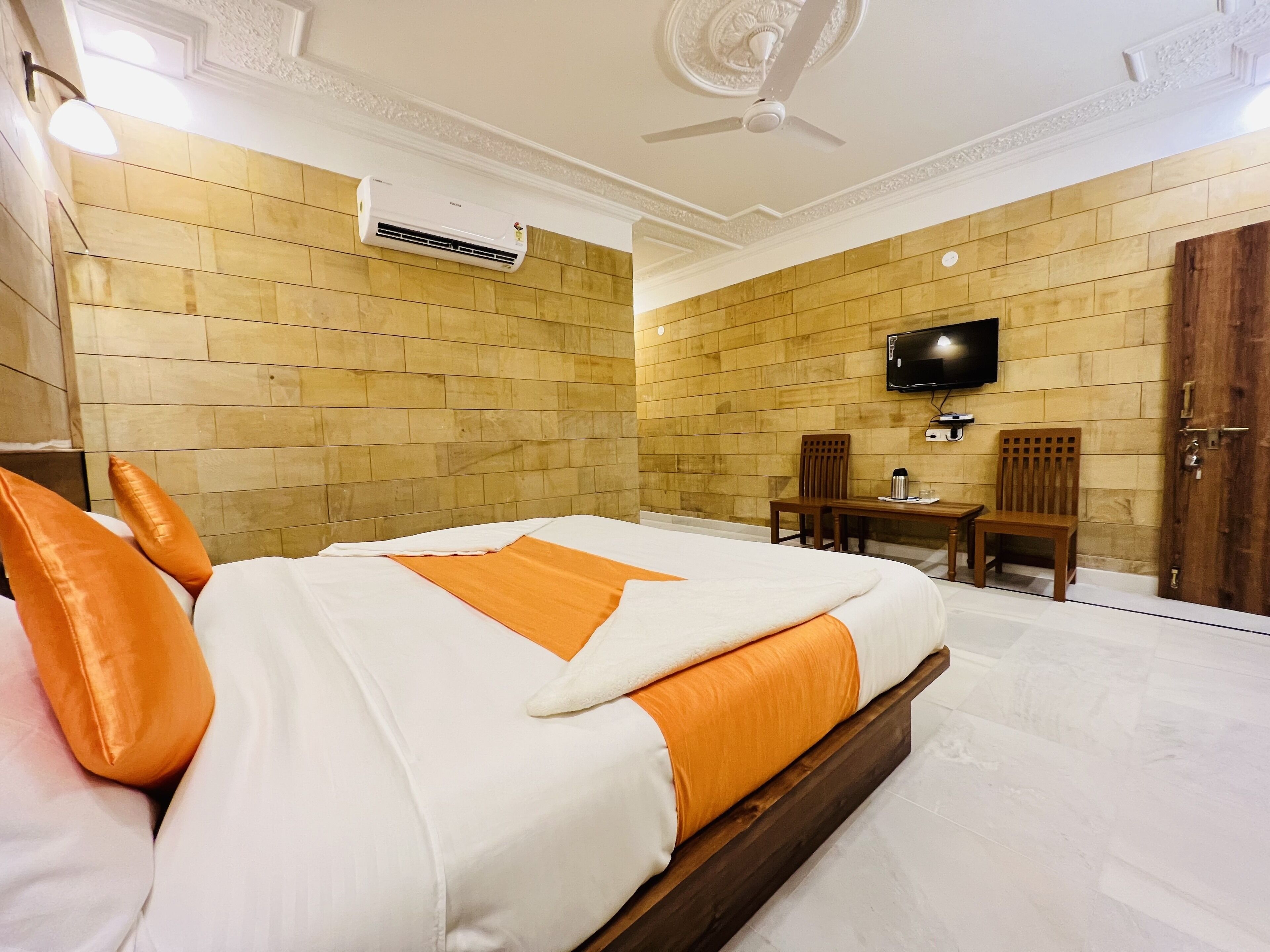 A hotel room with a large bed, a wooden headboard, a flat-screen TV mounted on the wall, a wooden table with chairs, and a wooden door.