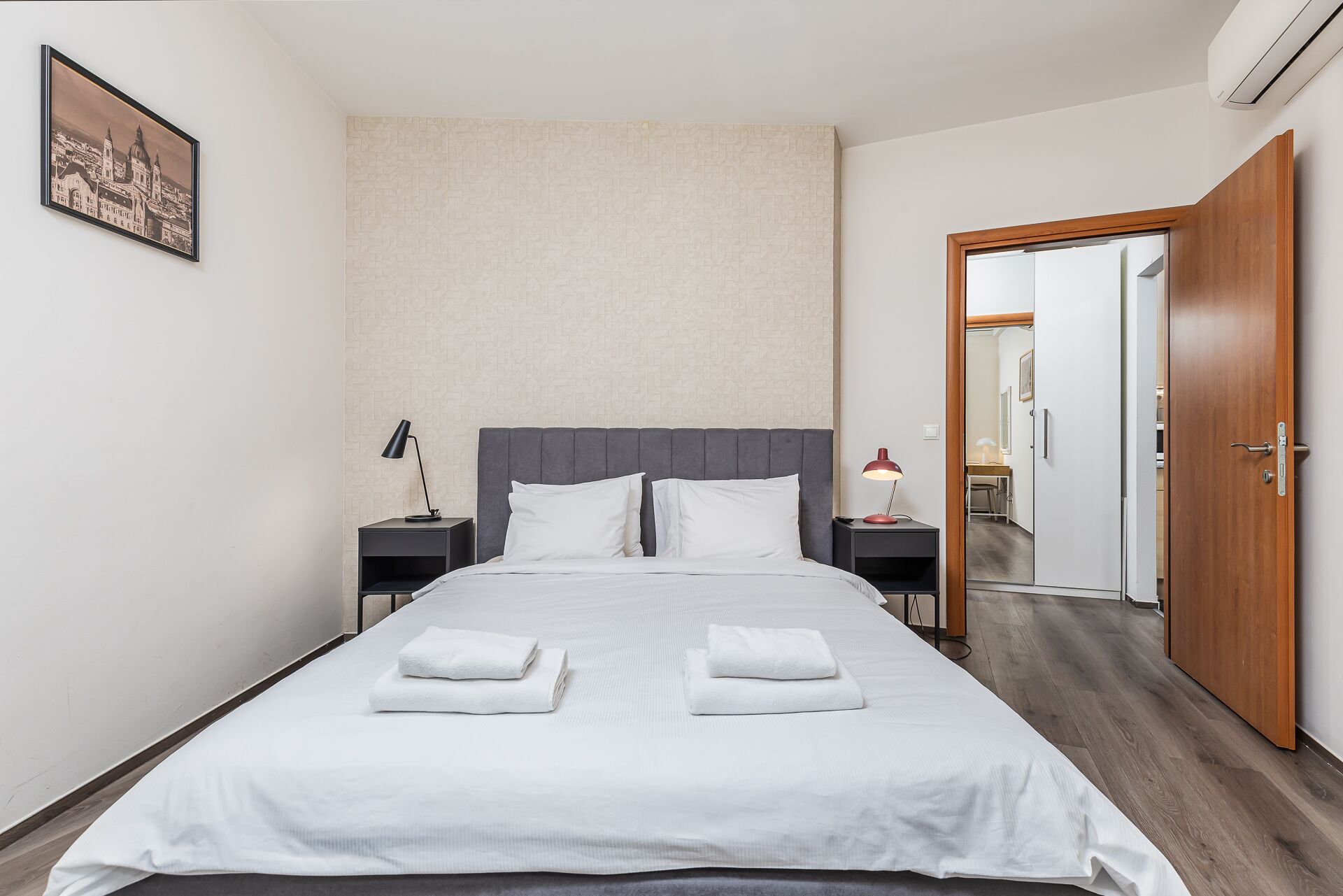Une chambre d’hôtel moderne avec un grand lit, deux tables de chevet, une petite table, un tableau encadré au mur et une porte en bois.