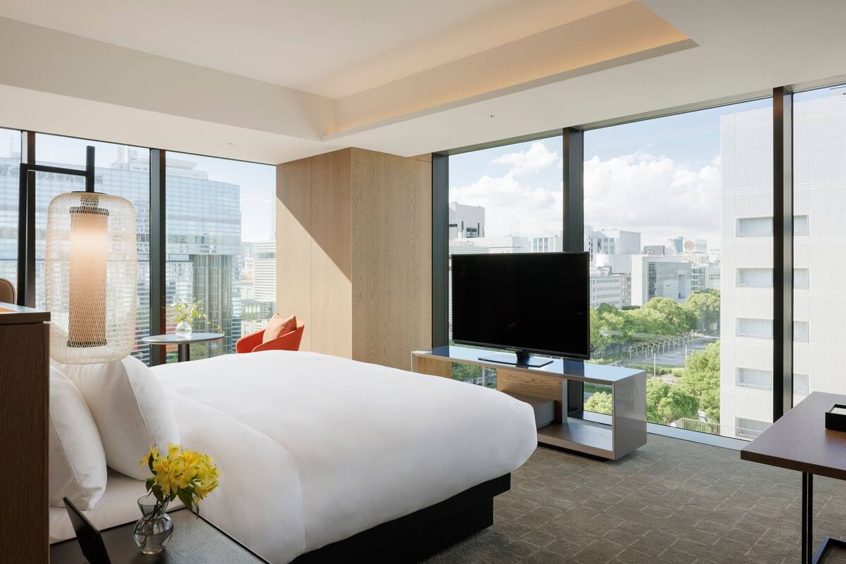 A modern hotel room with a large bed, a flat-screen TV, a city view, and a glass coffee table.