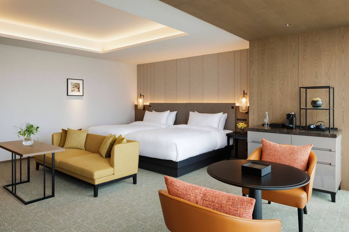 A modern hotel room with a large bed, a sofa, a dining table, and a kitchen area.