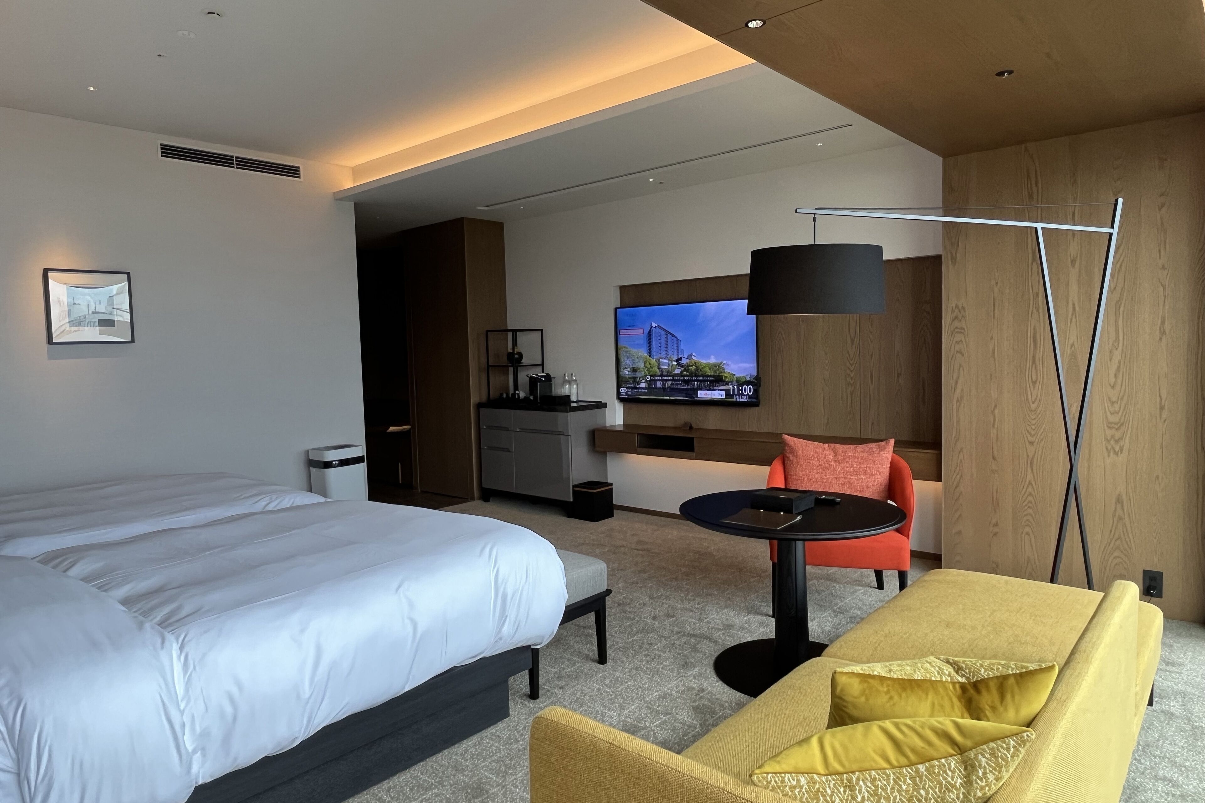 A modern hotel room with a large bed, a sofa, a small table, a TV, and a kitchenette.