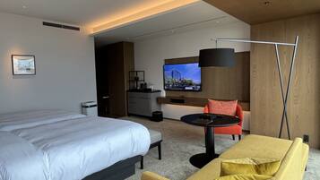 A modern hotel room with a large bed, a sofa, a small table, a TV, and a kitchenette.