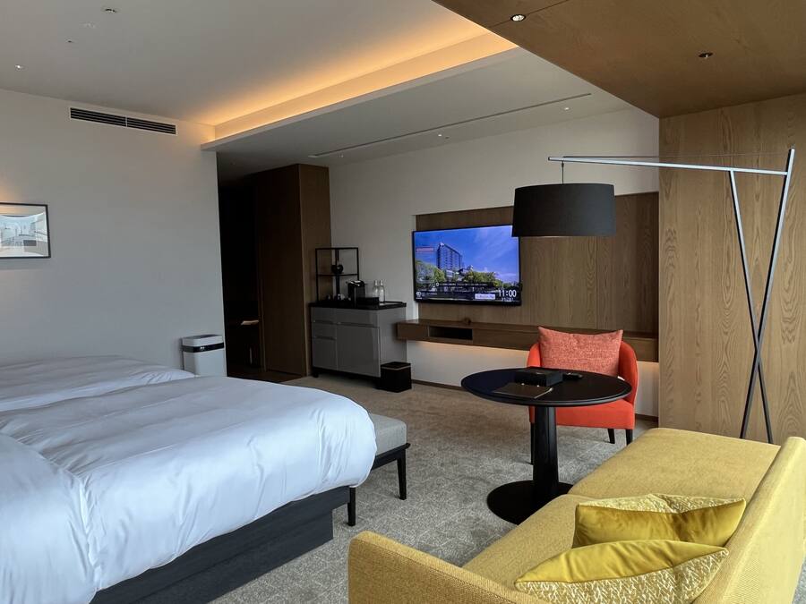 A modern hotel room with a large bed, a sofa, a small table, a TV, and a kitchenette.