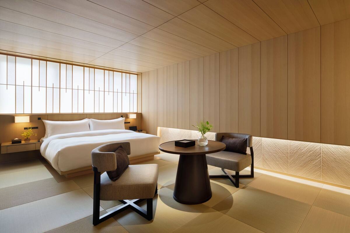 A modern hotel room with a large bed, two armchairs, a round table, and a view of the city through large windows.