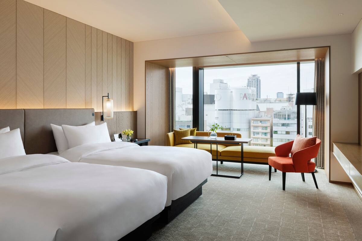 A hotel room with two beds, a sofa, a small table, and a chair. There is a large window with a view of buildings.