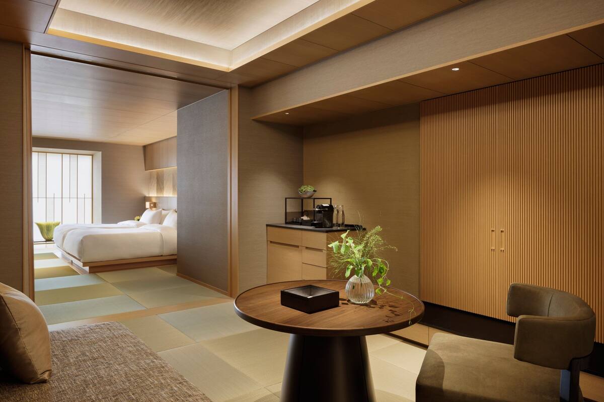 A modern hotel room with a bed, a desk, a chair, a round table, and a potted plant.