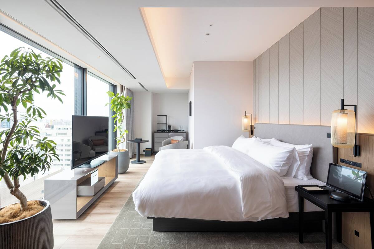 A modern hotel room with a large bed, a TV, a desk with a computer, and a large window with a city view.