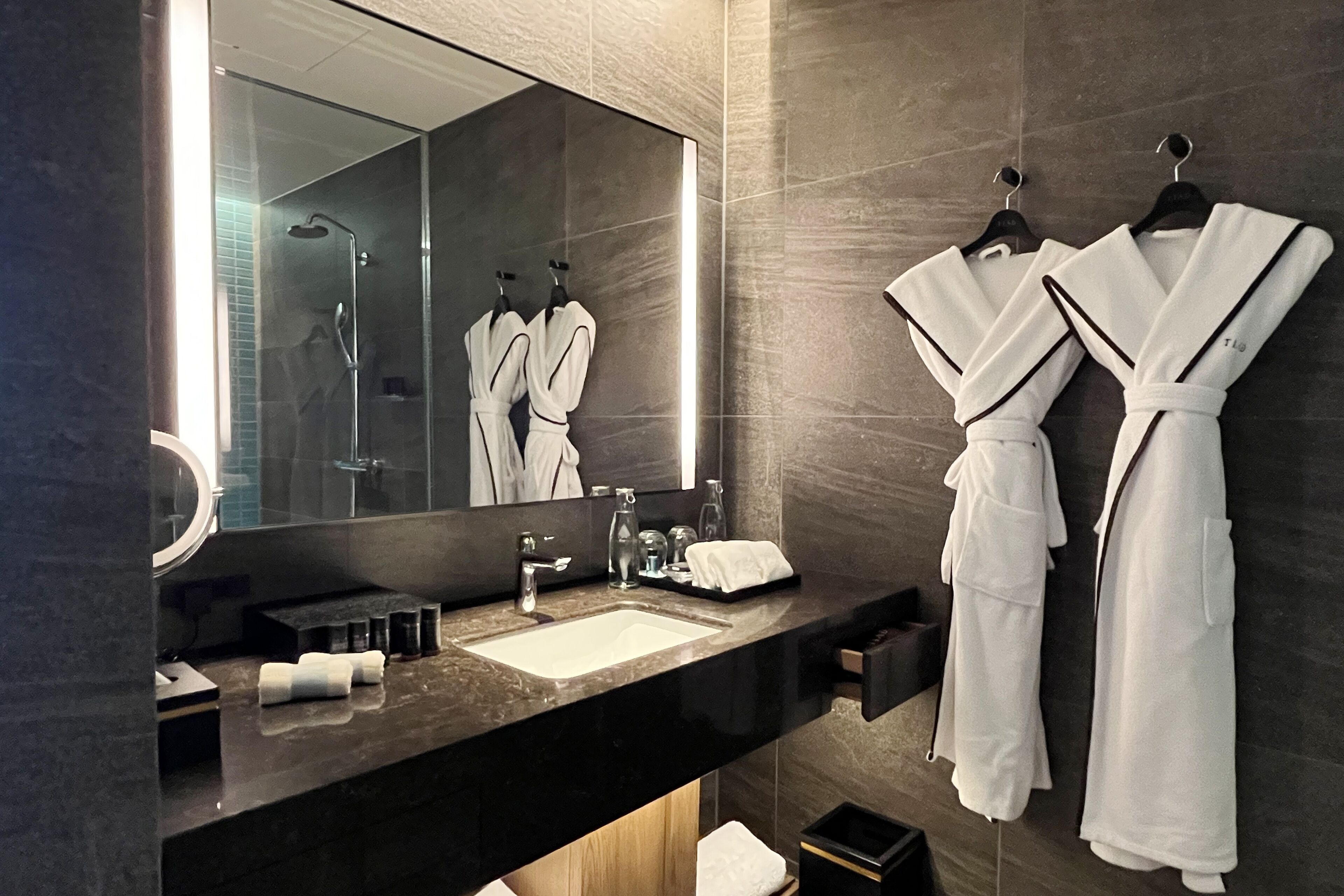 A modern bathroom with a large mirror, a sink, and two white robes hanging on hooks.