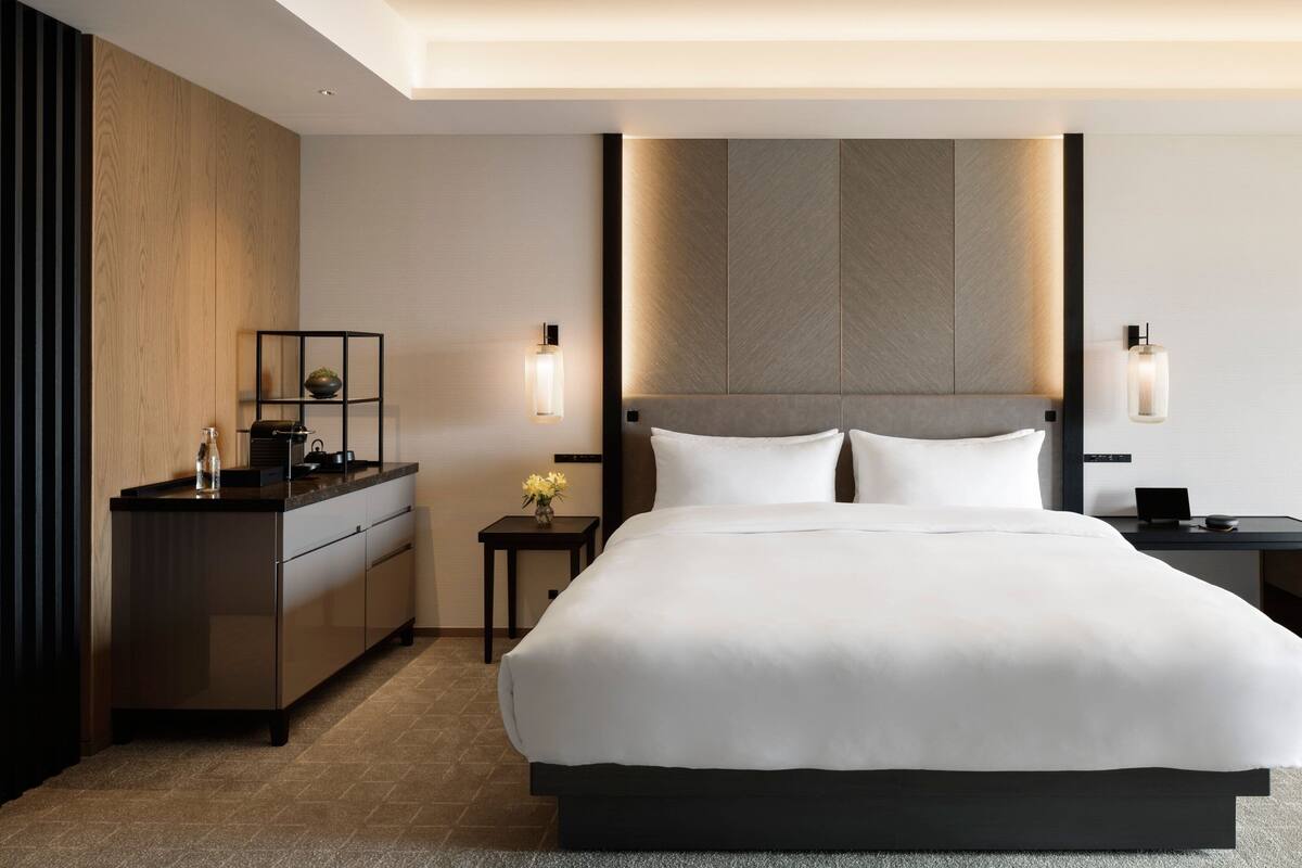 A modern hotel room with a large bed, a wooden sideboard, a small table with a vase, and two wall-mounted lamps.