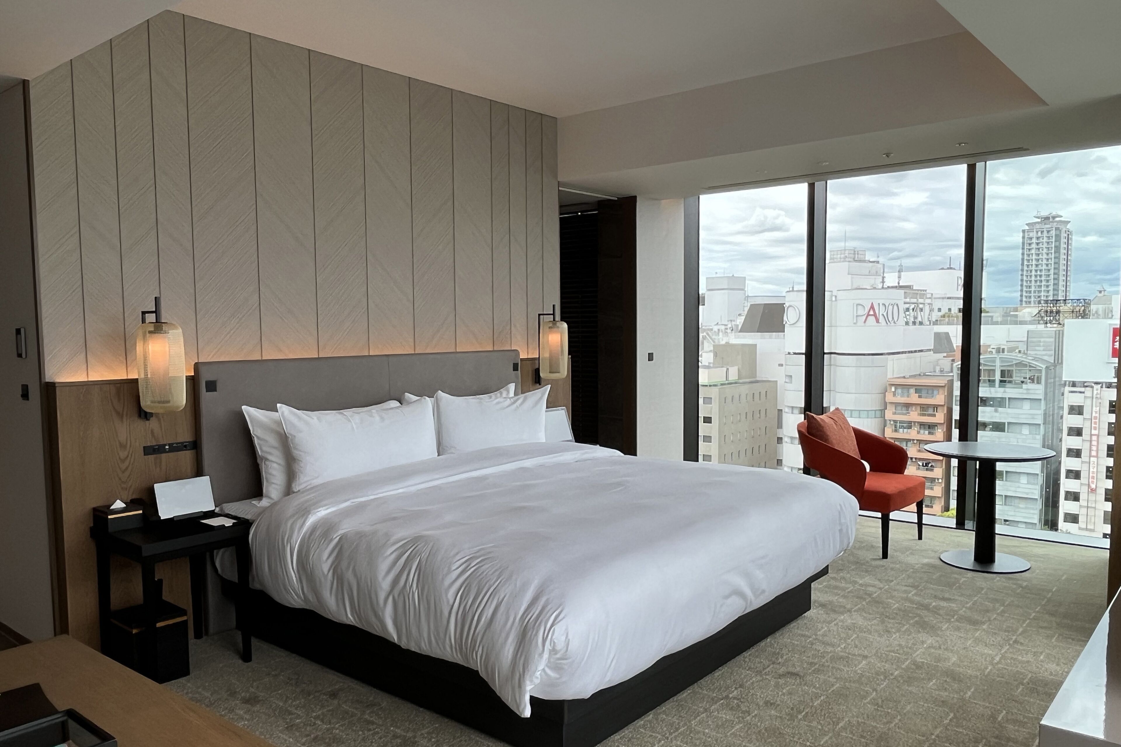 A modern hotel room with a large bed, a desk, a chair, and a view of the city through large windows.