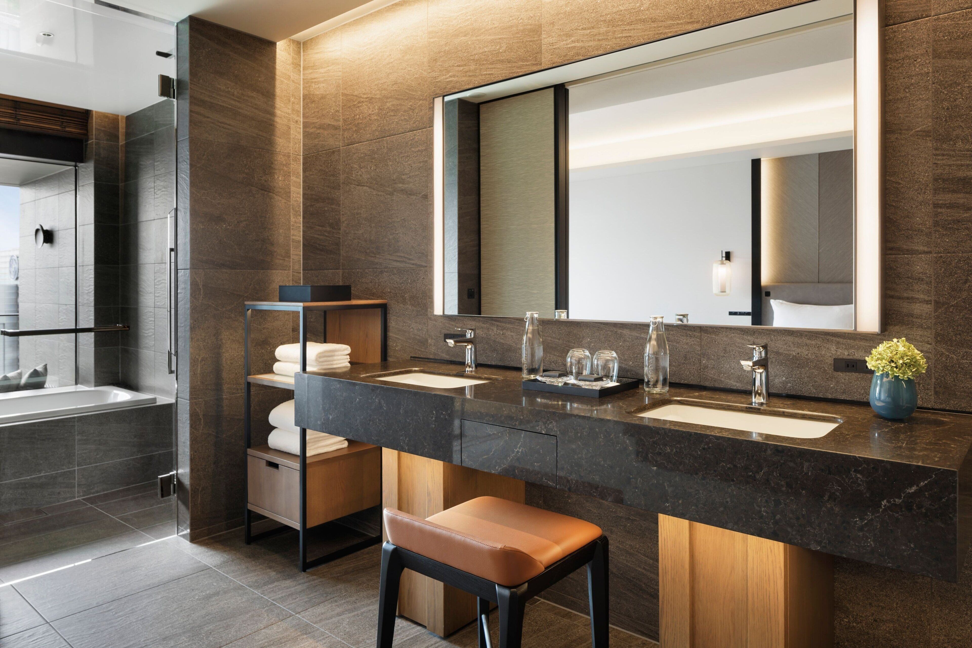 A modern bathroom with a large mirror, two sinks, and a shower. There are wooden cabinets and a bench.