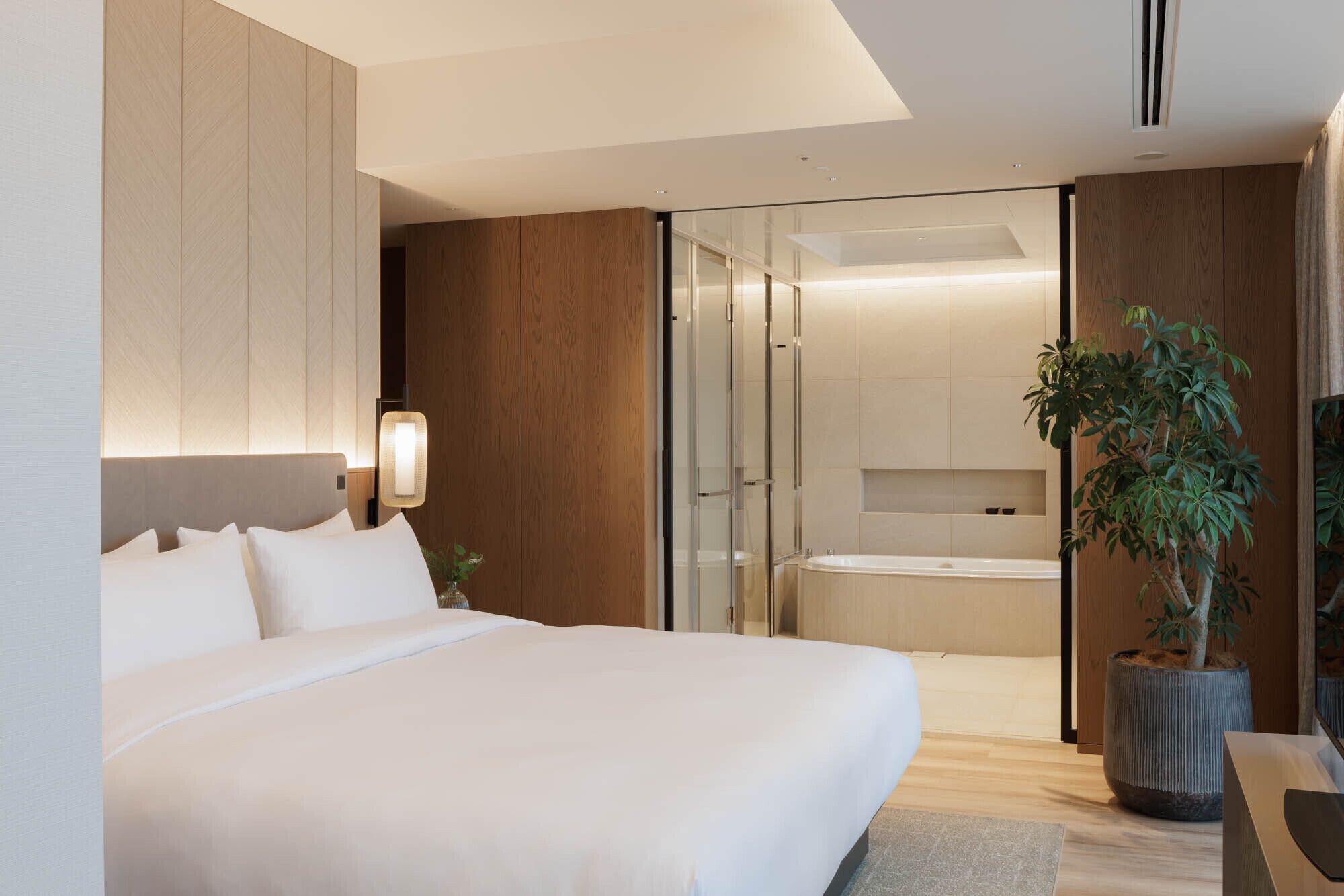 Quarto de hotel moderno com uma cama grande, box de chuveiro em vidro, banheira e um vaso de planta.