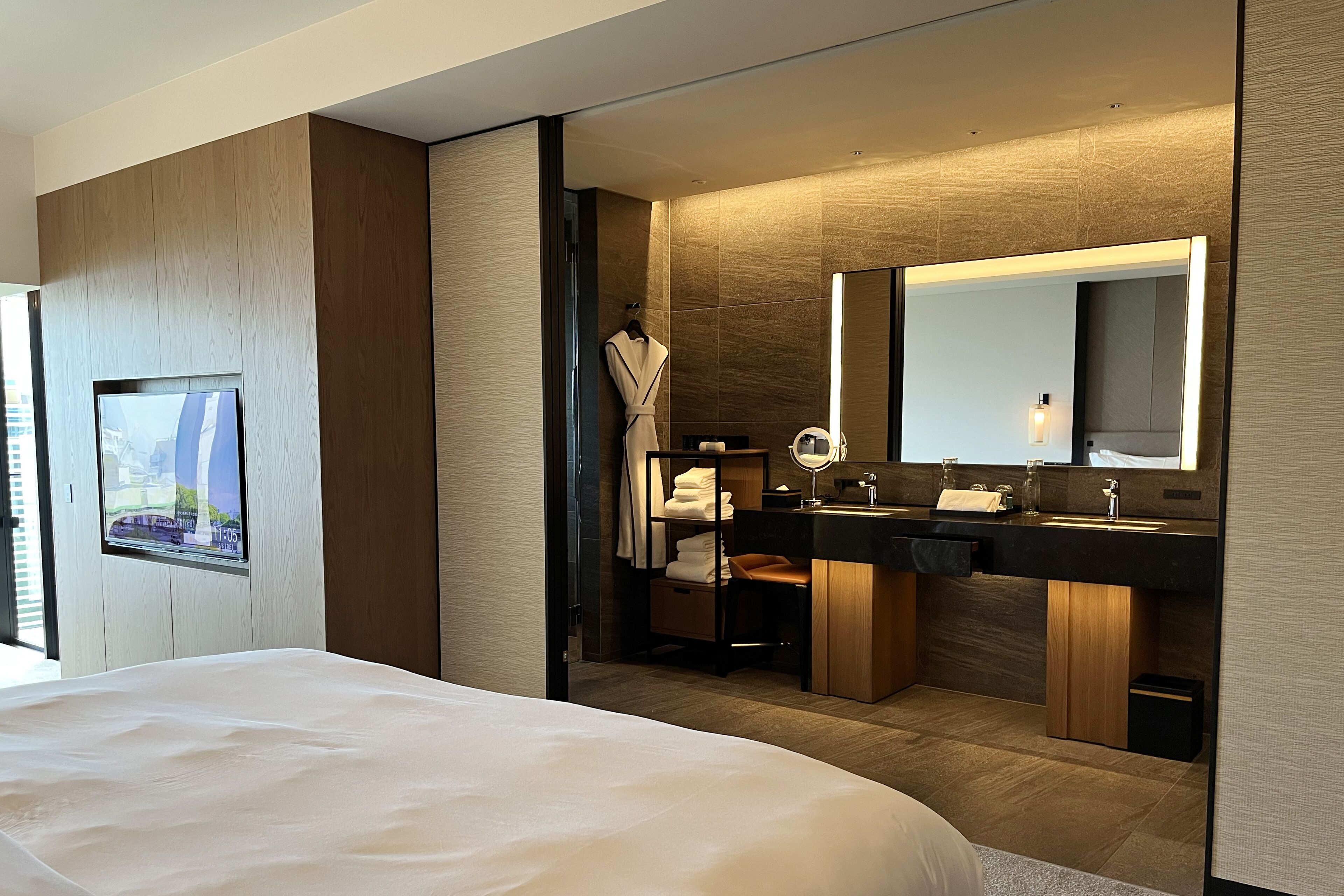 Una habitación de hotel moderna con una cama grande, un televisor montado en la pared, un baño con lavamanos dobles, un espejo y un perchero para bata.