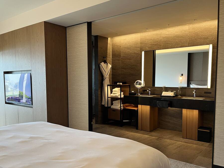 A modern hotel room with a large bed, a TV mounted on the wall, a bathroom with dual sinks, a mirror, and a robe rack.