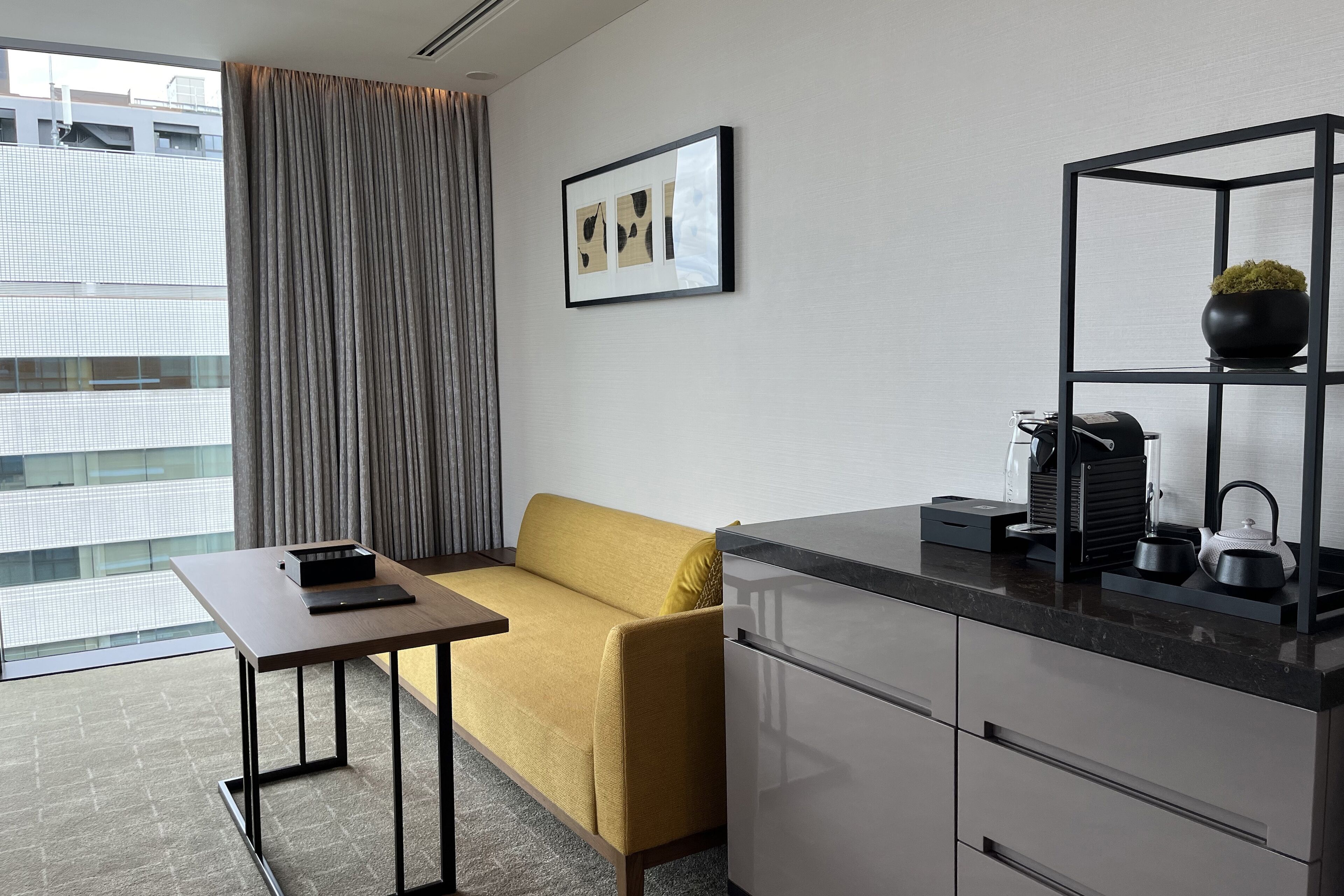Una habitación de hotel moderna con sofá, una mesita, una zona de cocina con cafetera y vistas a la ciudad.