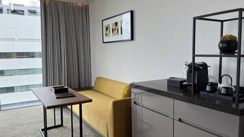 Una habitación de hotel moderna con sofá, una mesita, una zona de cocina con cafetera y vistas a la ciudad.