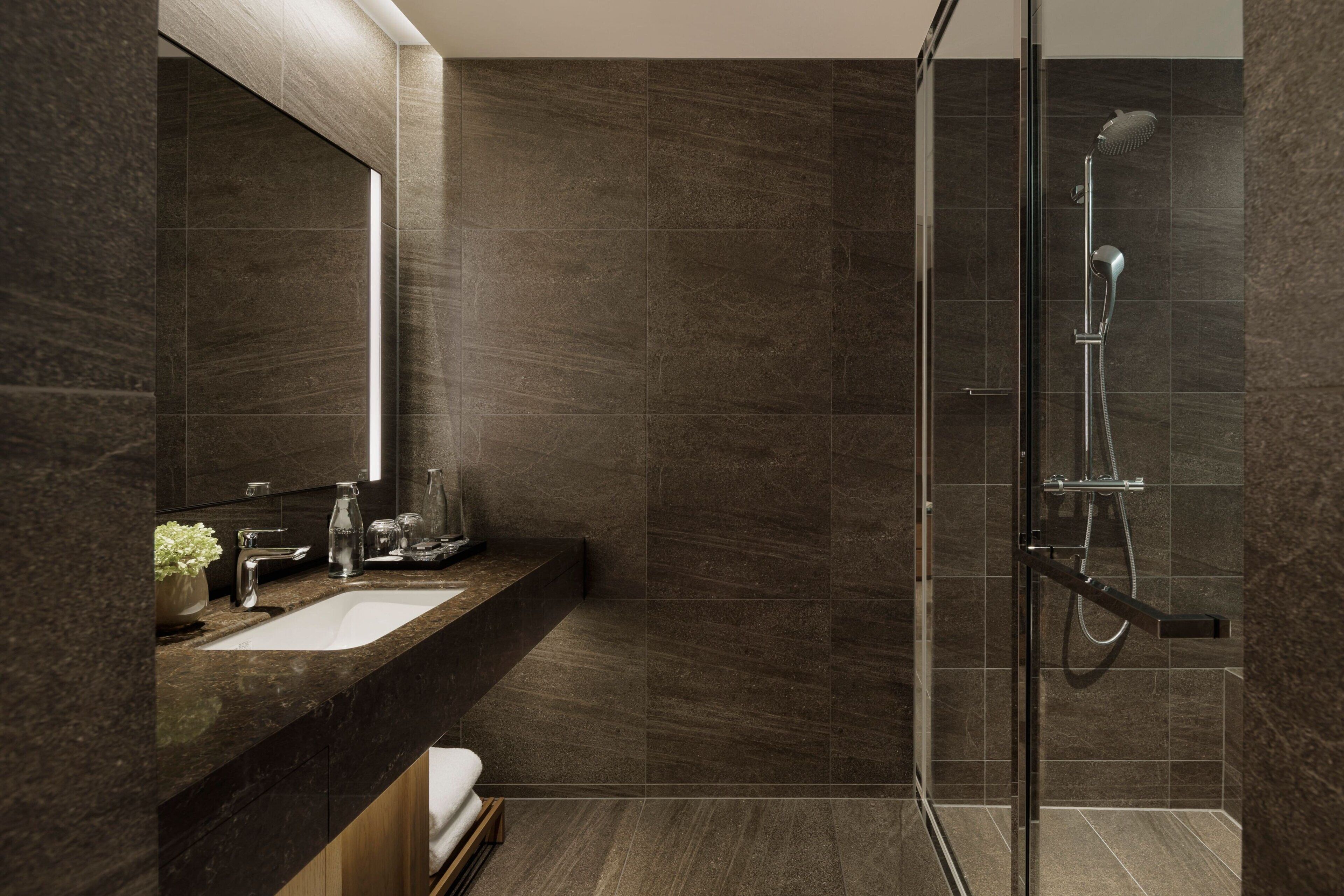 Un baño moderno con ducha envuelta en vidrio, paredes con azulejos oscuros, encimera de mármol y un espejo grande.