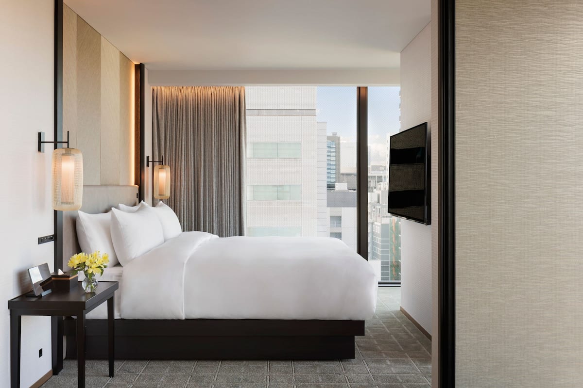 A modern hotel room with a large bed, a flat-screen TV, a nightstand with a lamp, and a view of the city through large windows.