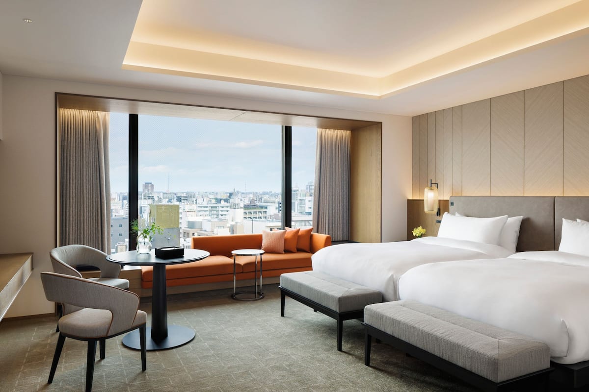 A hotel room with two beds, a sofa, a small table, and a chair. The room has large windows with curtains, a city view, and a modern design.