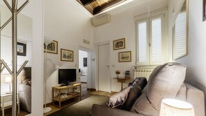 A living room with a sofa, TV, and a lamp. There are framed pictures on the wall and a window with blinds. - Minerva 5 - FromHometoRome (Roma)