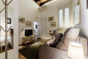 A living room with a sofa, TV, and a lamp. There are framed pictures on the wall and a window with blinds. - Minerva 5 - FromHometoRome (Roma)