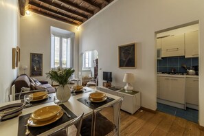 Een eetgedeelte met een tafel voor vier personen, een bank, een televisie, en een keuken zichtbaar door een open deur.