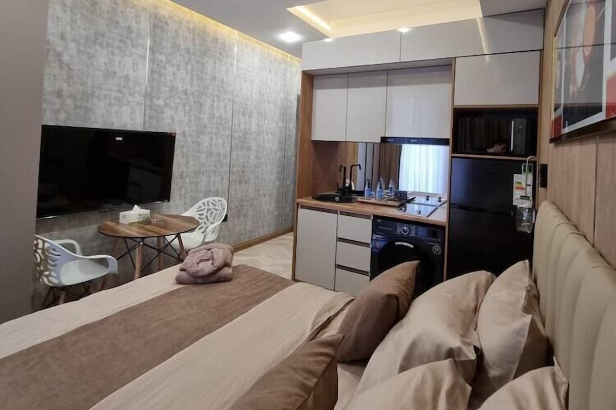 A modern hotel room with a bed, a TV, a small dining area, and a kitchenette.
