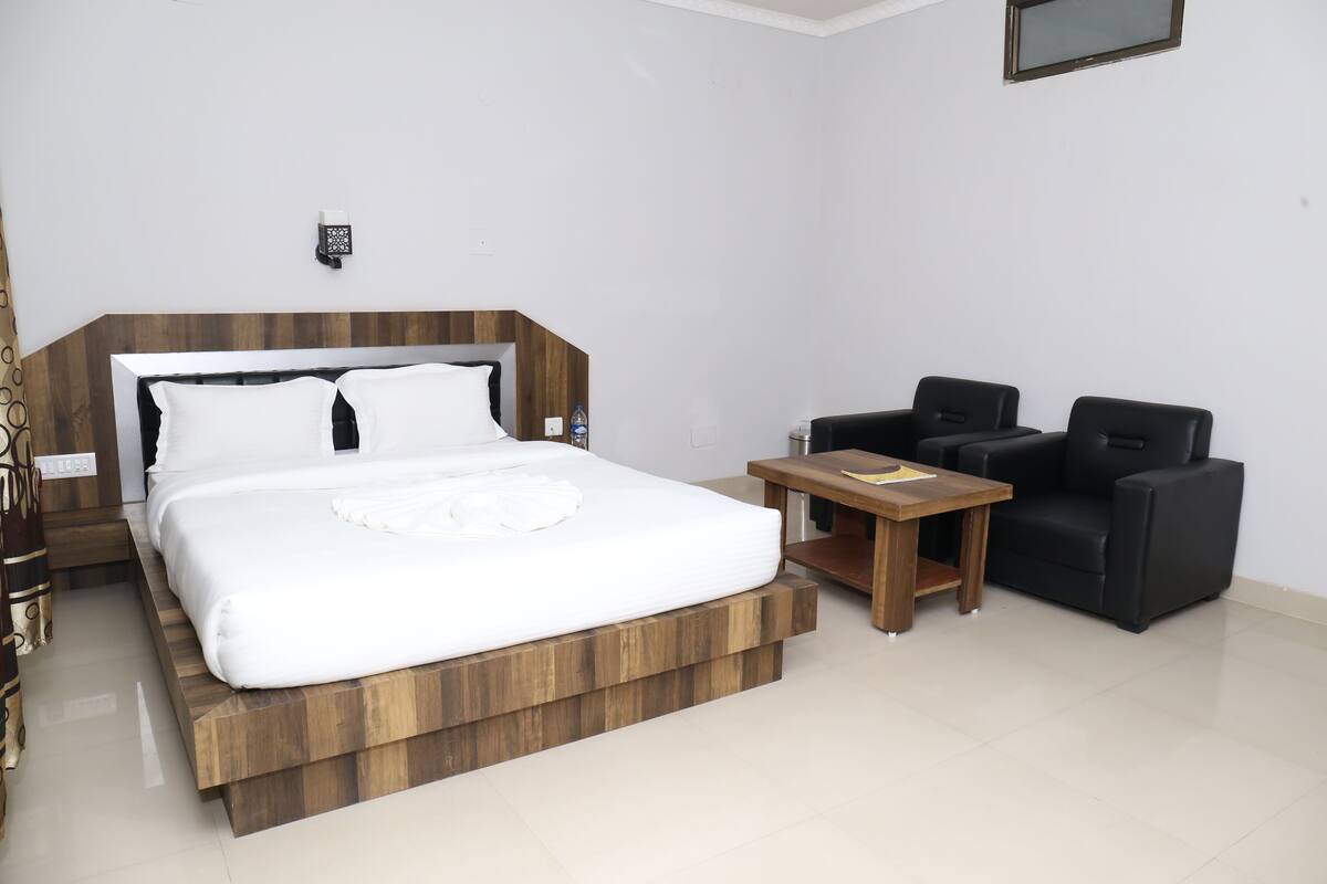 A modern hotel room with a large bed, a small wooden table, two black armchairs, and a wall-mounted light fixture.