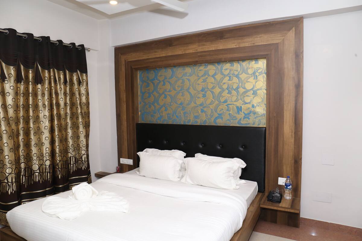 A neatly made bed with white linens and pillows, a wooden headboard with a decorative panel, a nightstand with a bottle and a phone, and a curtain with a pattern.