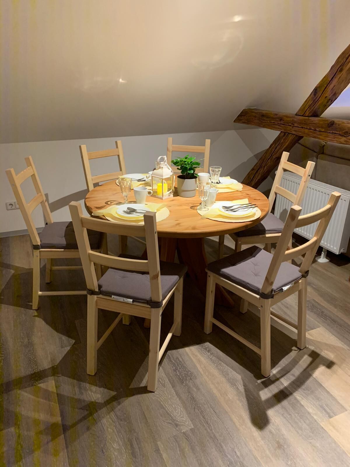 Une table de salle à manger ronde en bois, entourée de six chaises, chacune agrémentée d’un coussin. La table est mise avec des assiettes, des couverts, des verres et une petite plante en pot.