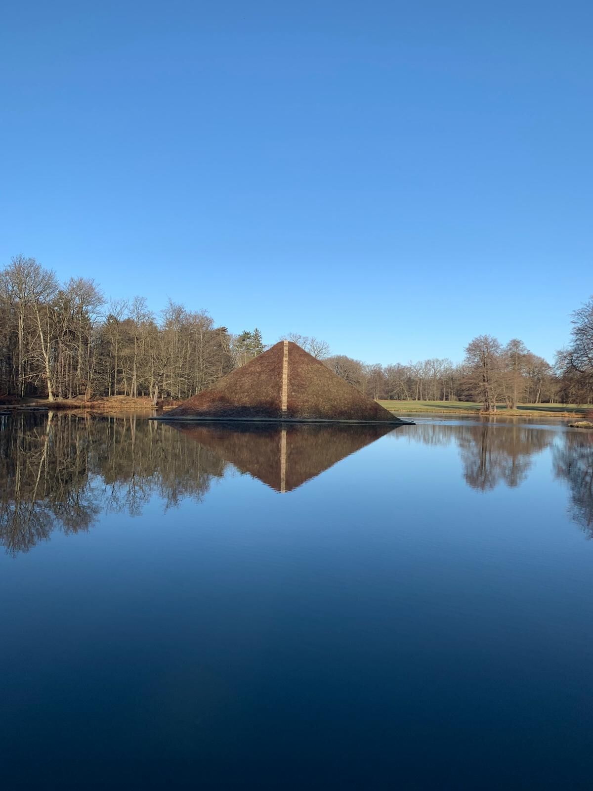 Un lac paisible, avec une structure en forme de pyramide au centre, entouré d’arbres et bordé d’un ciel dégagé.