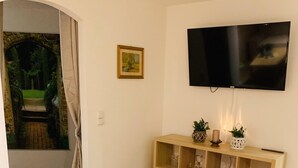 Una sala de estar con un televisor montado en la pared, un mueble de madera con objetos decorativos y un sofá color beige.